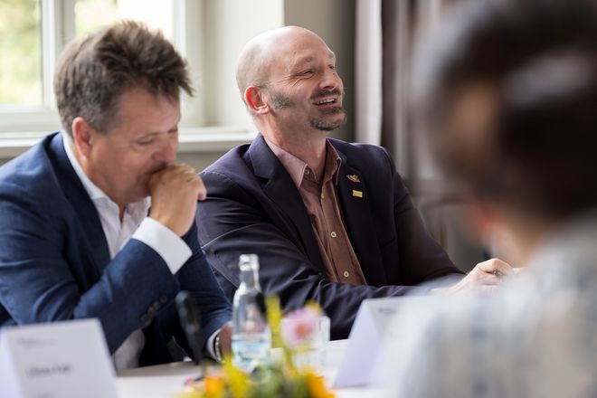 Simon Frisch und Jürgen Dusel nebeneinander am Besprechungstisch. Die Schärfe des Fotos liegt auf Frisch, der herzlich lacht. Dusel sitzt links neben ihm, er ist auf dem Foto unscharf gehalten, blickt schräg nach unten, eine Hand zur Faust vor dem Mund, als würde er sich räuspern. Vorne im Blick in der rechten Ecke ist eine weitere Person von hinten unscharf zu erkennen. Diese sitzt ihnen gegenüber.