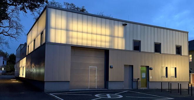 Außenansicht des Werkstattgebäudes bei Dämmerung, in dem die Werkstätten für Kunststoff, CNC, Modellbau und Holz untergebracht sind. Das zweigeschossige Gebäude mit transluzenter Fassade ist von innen beleuchtet; Fenster und Eingangsbereich sind sichtbar.