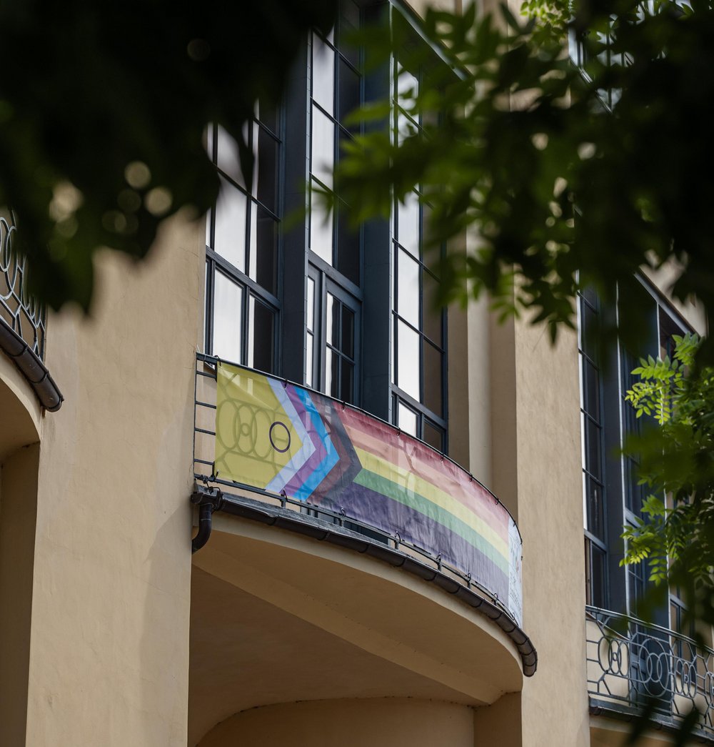 Das Bild zeigt ein Banner mit der »Inter*-inklusiven Progress-Pride-Flagge« am zentralen Balkon des Hauptgebäudes der Bauhaus-Universität Weimar.