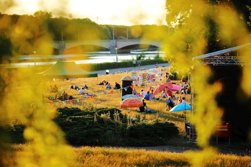 Menschen sitzen auf Picknick-Decken in der Abendsonne auf einer Wiese unter bunten Sonnenschirmen, Festivalgelände