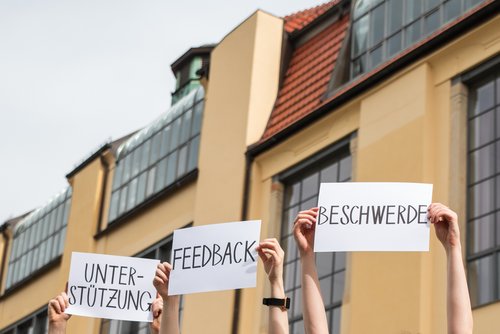 In front of the main building, hands hold up three pieces of paper. On them are the words: support, feedback, complaint