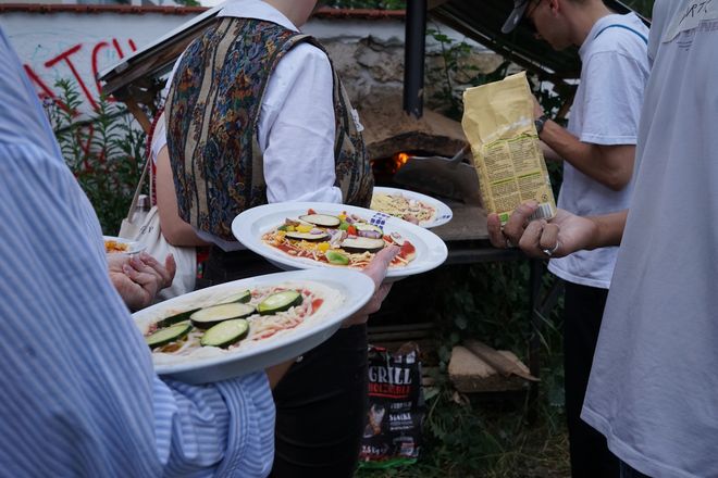 People wait in line with their topped pizzas in front of the pizza oven.