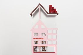 Modellhaftes Hausobjekt von Karla Zipfel in zarten Rosa- und Weißtönen, mit stilisierten Fenstern und dem Schriftzug »Modernisierung« an der Fassade.