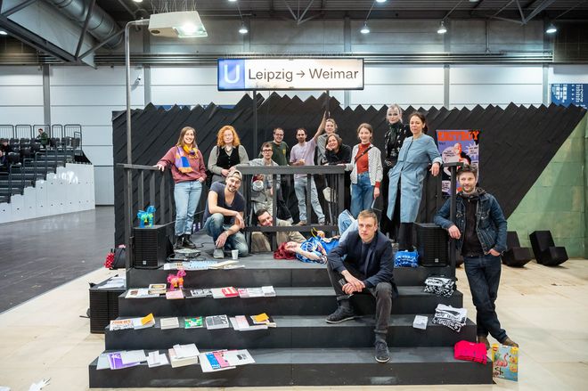 Group photo for »Ein Messestand für Leipzig«: several participants stand and sit on the Faculty of Art and Design’s trade fair stand, designed as a fictional underground station entrance. Above them is a sign reading »Leipzig → Weimar«.