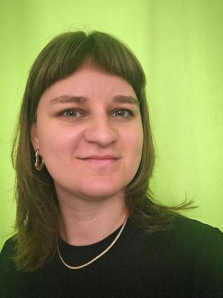 Porträt von Nele Seifert. Sie hat schulterlanges braunes Haar mit Pony und trägt goldfarbene Ohrringe sowie eine feine Halskette. Vor einem weich verlaufenden grünen Hintergrund blickt sie mit leichtem Lächeln in die Kamera.