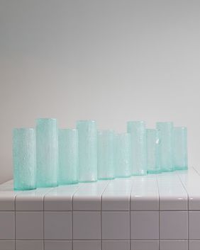 Row of translucent light blue glass vases of varying heights arranged on a white tiled surface. The vases are cylindrical and evenly spaced against a plain light gray background.