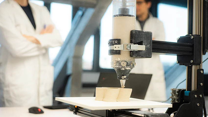 zwei Personen sehen dem 3D-Drucker bei der Arbeit zu