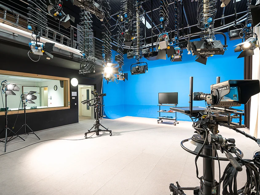 Innenaufnahme eines Videostudios: Im Vordergrund steht eine Videokamera auf einem Stativ, gerichtet auf einen Bereich mit weiterem Kamera- und Lichtequipment. Im Hintergrund sind Studiolampen, Mikrofone und weiteres technisches Zubehör für Videoaufnahmen erkennbar.