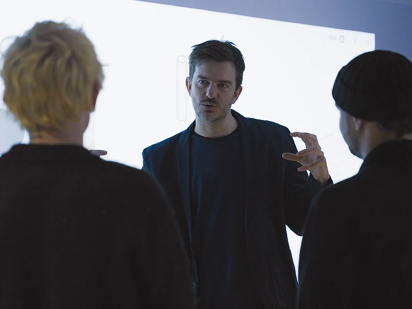 Prof. Thomas Pearce in conversation with students in front of a large-scale digital projection.