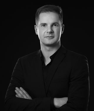 Portrait of Professor Jan Willmann against a black background. He wears a dark suit with a black shirt, stands with his arms crossed, and looks directly at the camera.