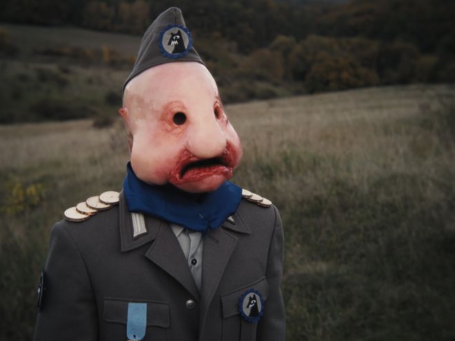 Nahaufnahme einer Person in Uniform mit glatter, puppenartiger Gesichtsform und runden Augenöffnungen.