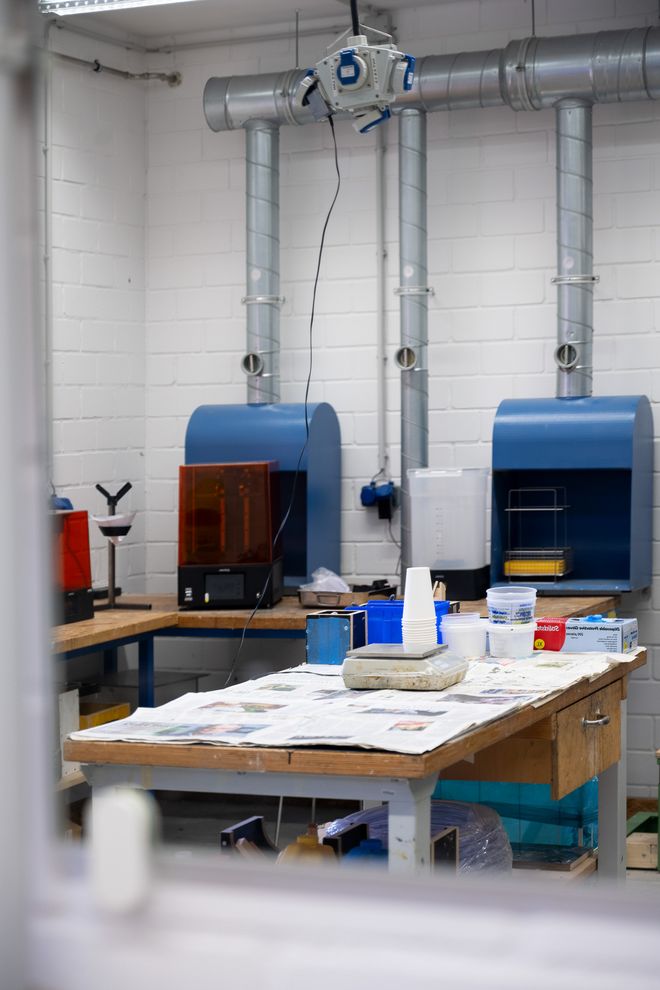 Arbeitsraum einer Kunststoffwerkstatt mit Werkbank, Abzugshauben, 3D-Druckern und Materialien auf einem mit Zeitungspapier bedeckten Tisch.