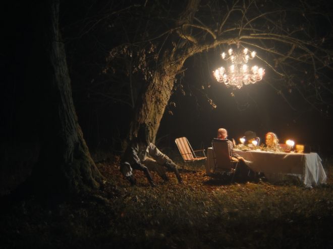 Mehrere Personen sitzen nachts im Wald an einem gedeckten Tisch unter einem hängenden Kronleuchter.