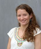 [Translate to English:] Porträtfoto von Dr. Elizabeth Watts vor einer grauen Betonwand: Sie hat lange, lockige, hellbraune Haare, braune Augen und trägt ein weißes, kurzärmeliges Oberteil mit buntem Muster am Kragen sowie ein Piercing im linken Nasenloch. Sie lächelt freundlich in die Kamera.