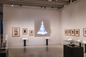 Ausstellungsansicht im Bauhaus-Museum Weimar: Grafische Arbeiten hängen gerahmt an der Wand, in der Mitte ist eine digitale Projektion einer abstrakten, skulpturalen Form zu sehen; Vitrinen mit Exponaten stehen im Raum.