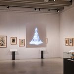 Ausstellungsansicht im Bauhaus-Museum Weimar: Grafische Arbeiten hängen gerahmt an der Wand, in der Mitte ist eine digitale Projektion einer abstrakten, skulpturalen Form zu sehen; Vitrinen mit Exponaten stehen im Raum.