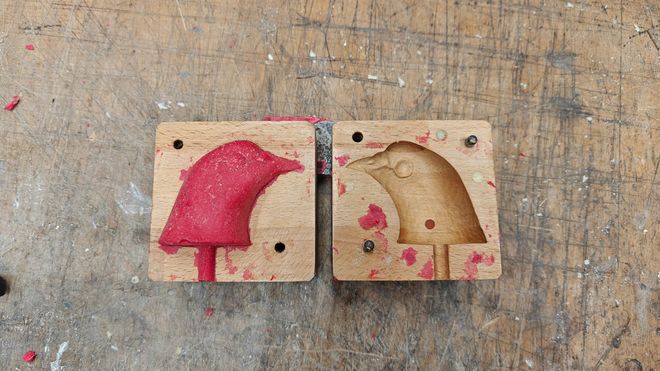 Zweiteilige Holzform mit eingelegter roter Masse auf einer stark beanspruchten Werkbank. Die Form zeigt zwei spiegelverkehrte Negativformen eines Vogelkopfes.