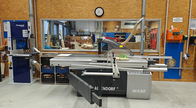 Große Formatkreissäge in einer Werkstatt, im Hintergrund weitere Maschinen und ein Einblick in angrenzende Arbeitsräume durch ein Fenster.