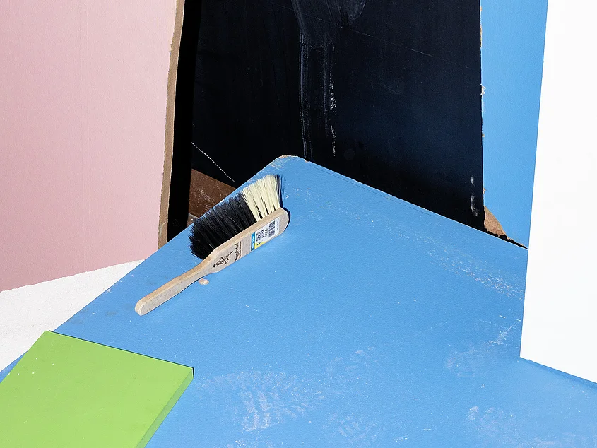 Detail of an artwork featuring coloured surfaces in blue, pink, green, black and white. A hand brush lies on the blue surface.