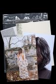 Still life with several printed magazine pages and photographs against a black background. Visible elements include a photograph of a person in front of rocks, a portrait with dark hair and a silver plastic sleeve.