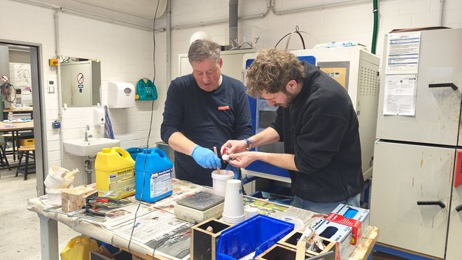 Zwei Personen arbeiten in einer Werkstatt an einem Tisch und mischen eine flüssige Masse in einem Kunststoffbecher. Auf dem Tisch stehen Kanister, Werkzeuge und Formen, der Arbeitsplatz ist mit Zeitungspapier abgedeckt.