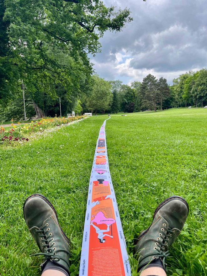 A long, colourfully printed fabric strip runs across a meadow in a park. A pair of shoes is visible in the foreground, while the illustrations and text on the strip lead into the distance.
