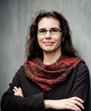 Portrait of Prof. Andrea Dreyer. Leaning against a concrete wall with her arms crossed, she looks into the camera with a slight smile. She wears glasses, a dark shirt, and a red patterned scarf.