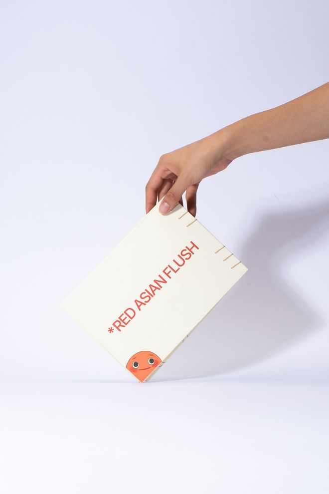 Product view of »RED AF* – Red Asian Flush« by Soo Ah Youn and Anastasia Fast: a hand holds a closed book titled »RED ASIAN FLUSH«. The illustrated character »Blush« peeks out from the lower edge.