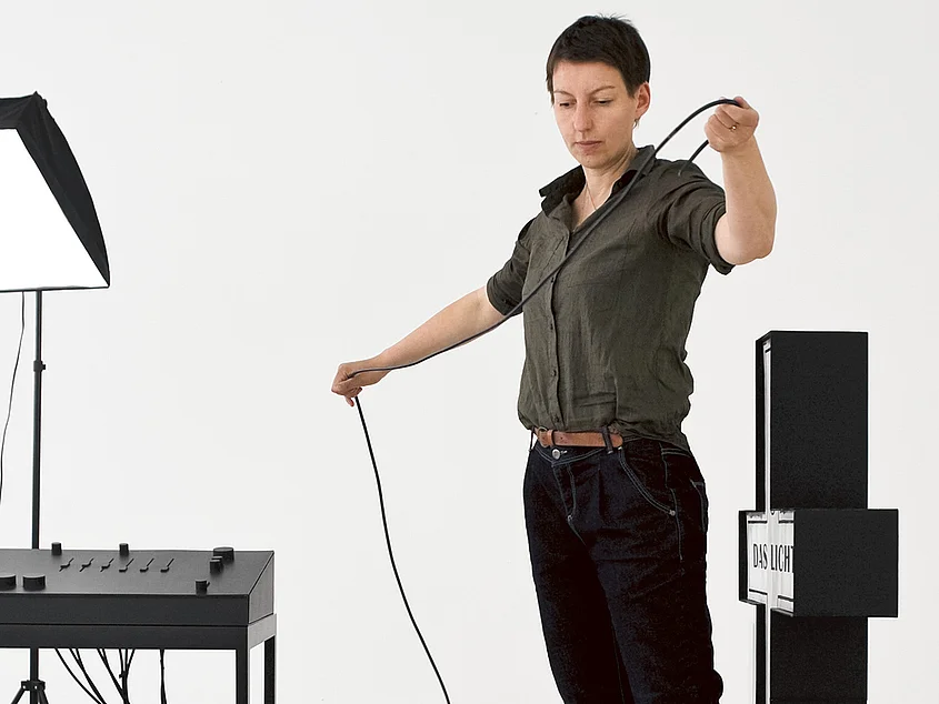 Portrait of Prof. Jana Gunstheimer. She holds a long black cable in her hands while standing among minimalist black objects and cables that are part of an installation in a bright studio.