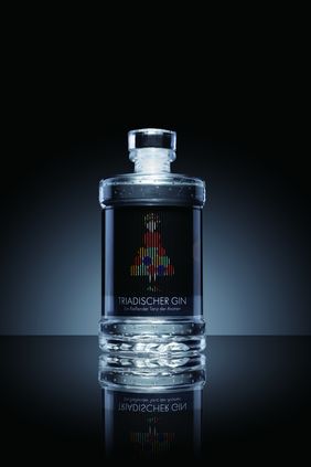 »TRIADISCHER GIN« by Xiaoyi Jin, Photo: Xiaoyi Jin Product photo of »TRIADISCHER GIN« by Xiaoyi Jin: a gin bottle stands against a dark green background on a reflective surface. The label shows an abstract, colourful figure referring to Oskar Schlemmer’s »Triadic Ballet«.