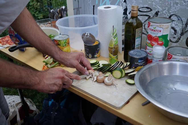 Two hands cut mushrooms and zucchinis on a wooden board. Other pizza ingredients are placed around the wooden board.