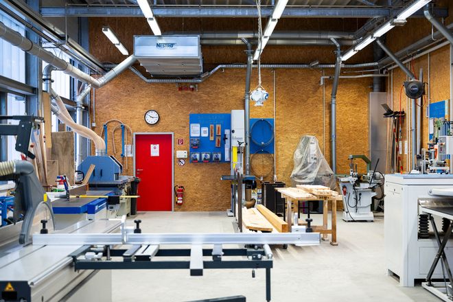 Large workshop room with various woodworking machines, workbenches, and extraction systems. A red door and technical connections are visible on the wall.
