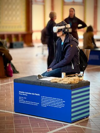 Person sitzt mit VR-Brille an einer Präsentationsstation und erlebt den 360-Grad-Film »Arbeiter betreten die Fabrik« von Florian Wehking.