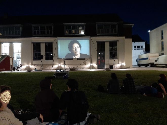 Photo during the movie screening. It is dark. In the foreground, guests are sitting on the lawn and looking at the screen in the background.
