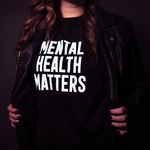 The photo shows the torso of a woman. She She is wearing a black T-shirt with the white inscription »Mental Health Matters«.