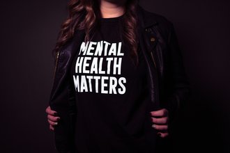 The photo shows the torso of a woman. She She is wearing a black T-shirt with the white inscription »Mental Health Matters«.