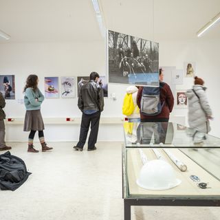 Exhibition space with several visitors viewing photographs and graphic works displayed on white walls. In the foreground, a glass display case contains objects, including a white safety helmet; additional exhibits are suspended in the room.
