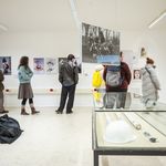 Exhibition space with several visitors viewing photographs and graphic works displayed on white walls. In the foreground, a glass display case contains objects, including a white safety helmet; additional exhibits are suspended in the room.