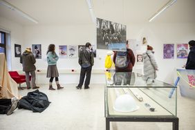 Foto: Thomas Müller Ausstellungsraum mit mehreren Besucher*innen, die Fotografien und grafische Arbeiten an weißen Wänden betrachten. Im Vordergrund steht eine Glasvitrine mit Objekten, darunter ein weißer Schutzhelm; weitere Exponate hängen frei im Raum.