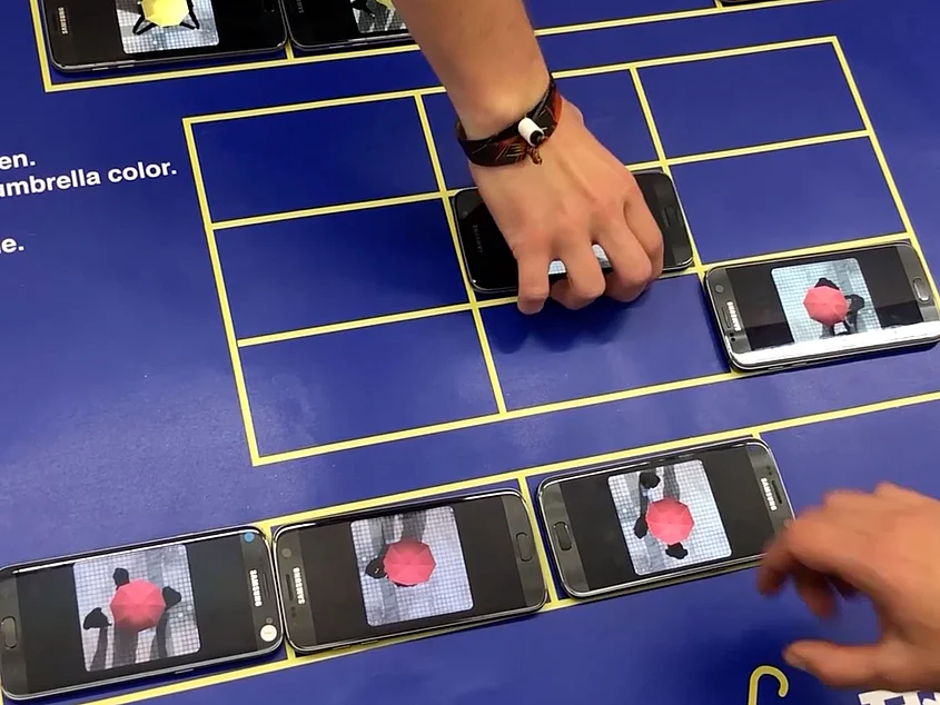 Mehrere Smartphones liegen auf einem Spielfeld mit gelbem Raster. Auf den Displays sind rote und gelbe Figuren zu sehen; zwei Hände bewegen die Geräte.