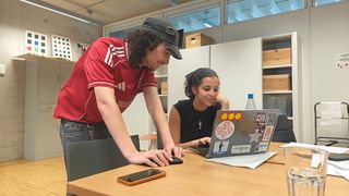 Zwei Studierende arbeiten gemeinsam an einem Laptop. Einer steht über den Tisch gebeugt, die andere sitzt vor dem Rechner.