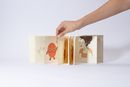 Book view of »RED AF* – Red Asian Flush« by Soo Ah Youn and Anastasia Fast: a leporello is unfolded, showing several illustrated scenes with the character »Blush« and a protagonist.