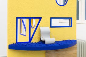 Detail eines architektonischen Objektmodells von Karla Zipfel mit gelber Wand, blauen Fensterrahmen und einer kleinen Sitzskulptur auf einer strukturierten Plattform.