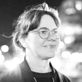 Schwarz-Weiß-Porträt von Barbara Kotte mit Brille. Sie blickt leicht zur Seite, während unscharfe Lichter im Hintergrund eine nächtliche Atmosphäre erzeugen.