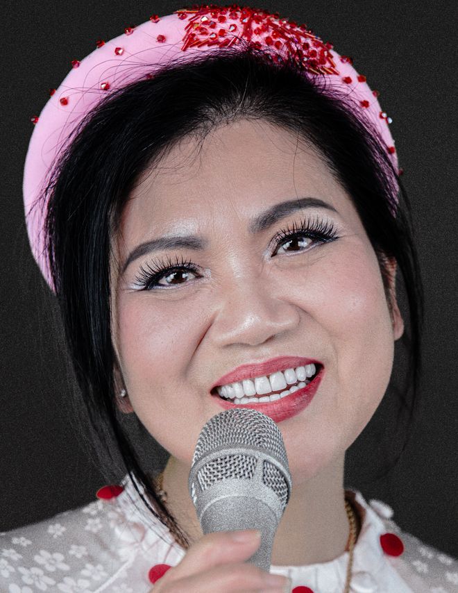 Portrait of a smiling woman holding a microphone. She wears a white top with red dots and a pink headband while looking into the camera.