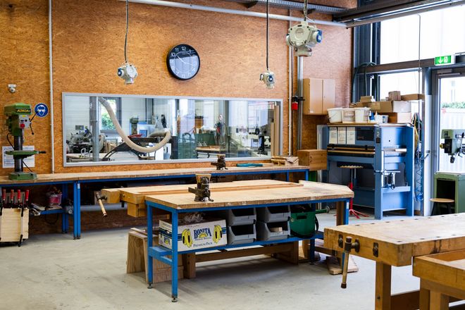 Workshop room with several sturdy wooden workbenches, machines and tools. Suction hoses, a clock and technical installations hang on the wall.