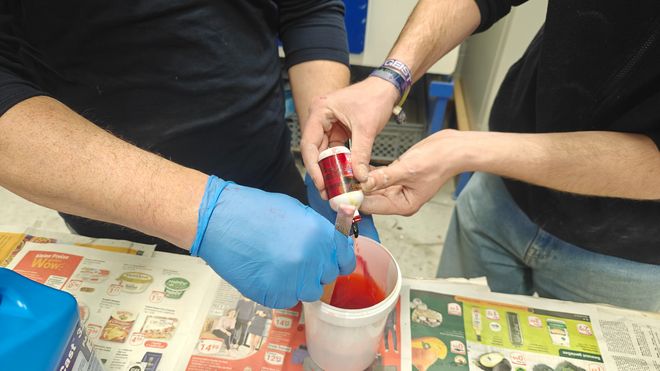 Detailaufnahme von zwei Paar Händen beim Einfärben einer flüssigen Masse. Eine rote Farbflüssigkeit wird aus einer kleinen Flasche in einen Becher gegossen und mit einem Spatel verrührt.