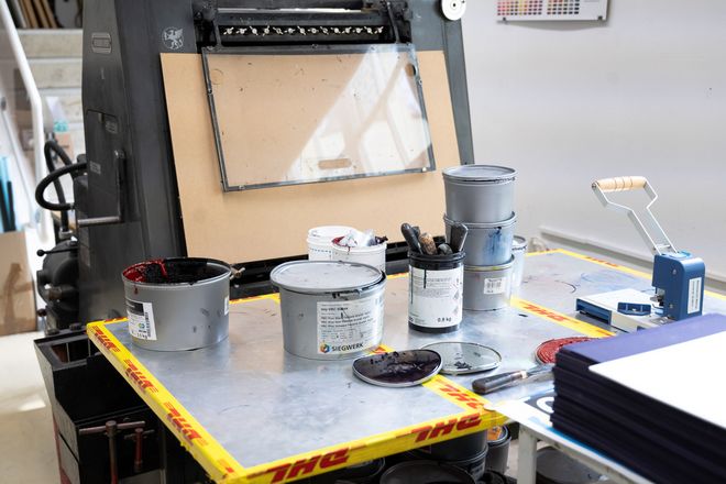 Arbeitstisch mit geöffneten Farbdosen, Spachteln und Druckfarbe vor einer Druckpresse.