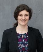 Porträtfoto von Miriam Benteler vor einer grauen Betonwand: Sie hat lange, lockige, braune Haare, die Zu einem Pferdeschwanz gebunden sind. Sie trägt eine dunkelblaues Oberteil mit rotem und weißem Blumenmuster und darüber einen schwarzen Blazer. Sie lächelt freundlich in die Kamera. 