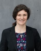Portrait photo of Miriam Benteler. She has brown, curly hair tied back in a ponytail. She is wearing a dark-blue top with red and white flowers on it, a black jacket, and she is smiling at the camera.